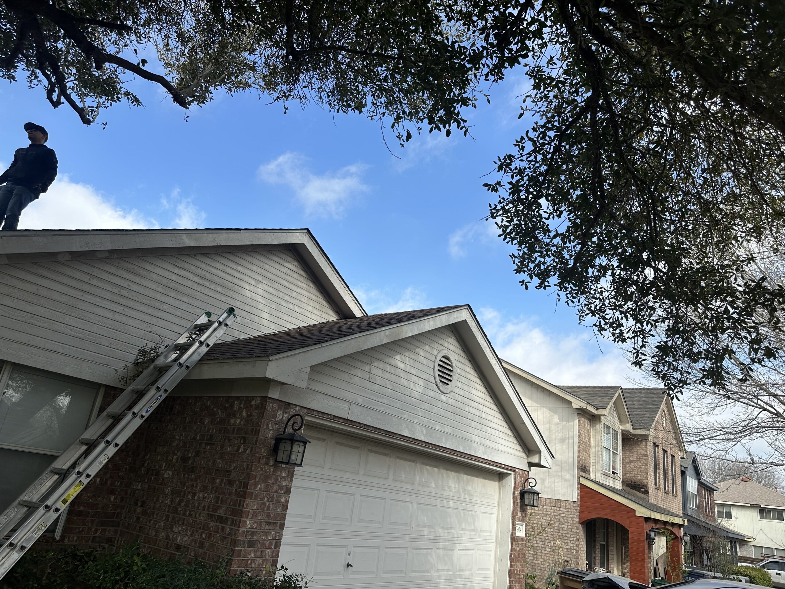 A person stands on the edge of a house roof near a ladder, with neighboring houses and tree branches visible under a partly cloudy sky.